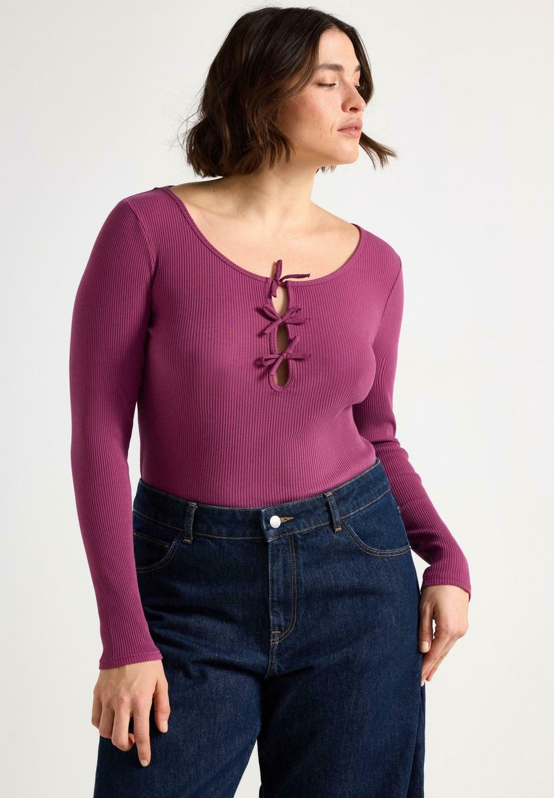 Kvinna som bär en långärmad lila topp med ribbad struktur och tre knutna rosetter på bröstet, kombinerad med mörkblå höga jeans, poserar i profil.