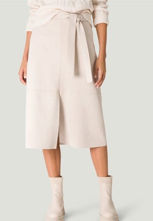 Frau trägt einen beigen Midirock mit Frontschlitz und gebundenem Gürtel, kombiniert mit cremefarbenen groben Stiefeletten und einem strukturierten Pullover.