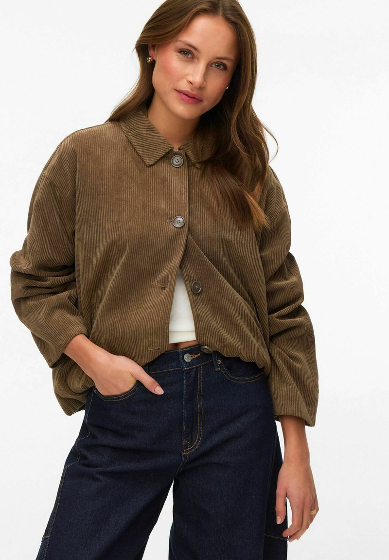 Nainen pukeutuneena ruskeaan puuvillaflanelitakkiseen napitettavaan takkiin valkoisen paidan päällä ja tummansinisiin korkeavyötäröisiin farkkuihin, toinen käsi taskussa, poseeraa valkoista taustaa vasten.