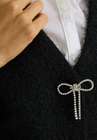 Un pull noir en peluche avec un décolleté en V profond, orné d'une broche en forme de nœud en rhinestones, décorée de pierres claires et de deux longues attaches.
