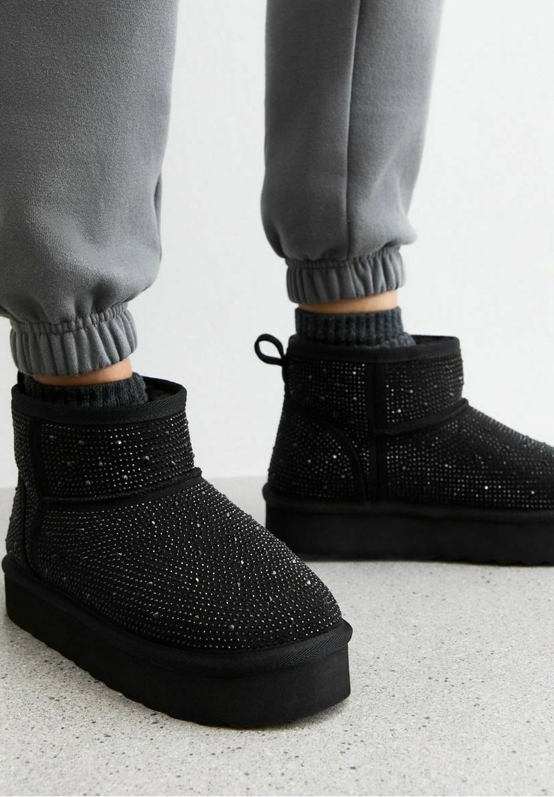 Botas negras de tobillo cubiertas de pequeños rhinestones, con suela plana y una lengüeta en la parte posterior, combinadas con pantalones de chándal grises con puños elásticos.