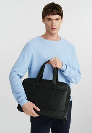 Młody mężczyzna w jasnoniebieskim swetrze trzymający czarną skórzaną teczkę z podwójnymi uchwytami na białym tle.