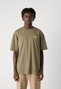Olivenfarvet oversized t-shirt i bomuld, med et lille gult logo på venstre brystområde og korte ærmer. Simpelt design.