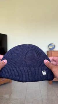 Gorro de malha azul-marinho com textura canelada, apresentando um pequeno logótipo quadrado. O chapéu é segurado com ambas as mãos, mostrando a sua forma e design.