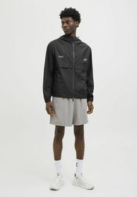 Zwarte lichtgewicht herenjack met rits, verstelbare capuchon en reflecterend logo. Gecombineerd met grijze shorts en witte sneakers.