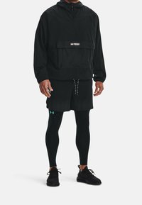 Giacca leggera nera con tasca frontale, abbinata a pantaloni corti neri e leggings neri. Presenta un logo del marchio turchese sui pantaloni corti.