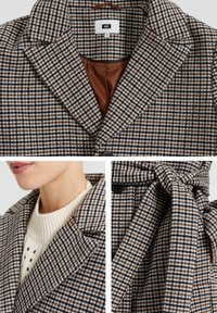 Brun och vit hundtandsmönstrad kappa med slogkrage, knappdetaljer och bälte av tyg. Insidan har en brun foder.