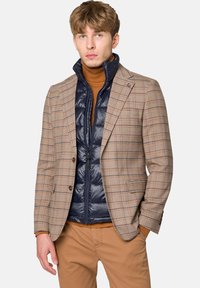 Kratkowana marynarka w beżowym i brązowym kolorze, nałożona na granatową kamizelkę puchową, z prążkowanym golfem. Kombinację uzupełniają jasnobeżowe spodnie.