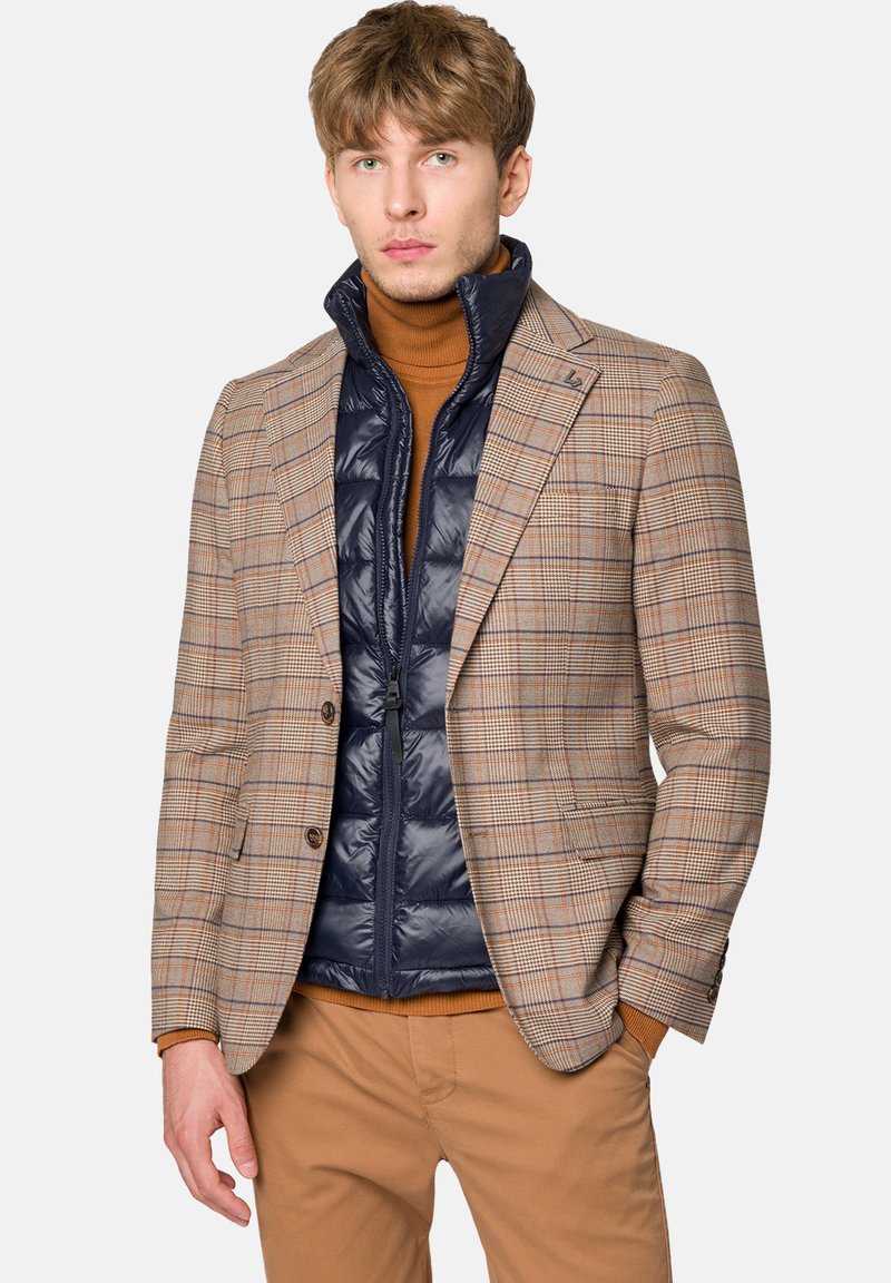 Kratkowana marynarka w beżowym i brązowym kolorze, nałożona na granatową kamizelkę puchową, z prążkowanym golfem. Kombinację uzupełniają jasnobeżowe spodnie.