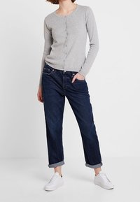 Harmaan pitkämielinen neuletakki, jossa on napit, yhdistettynä käännettyihin tummansinisiin farkkuihin. Valkoiset tennarit täydentävät rentoa asua.