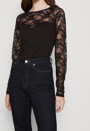 Vrouw draagt een zwarte kanten top met lange mouwen en bloemenpatroon, gecombineerd met een donkerblauwe high-waisted jeans.