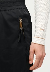 Κοντινό πλάνο χεριού δίπλα σε μαύρο παντελόνι με χρυσό φερμουάρ και δερμάτινο τραβηχτήρι, που φοράει λευκό μανίκι με καπιτονέ σχέδιο, μπεζ ραφές και μανσέτα.