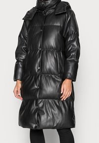 Černý prošívaný kabát z umělé kůže s vysokým límcem, patentními knoflíky a volným, nadměrným střihem. Hladký povrch a prošívaný vzor.