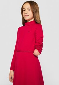 Piros, hosszú ujjas ruha, álló nyakkal, díszítő övvel. Az anyag lágy hatású; feltűnő logó látható a mellkason.