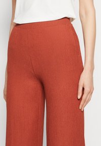 Gros plan d'une personne portant un pantalon large taille haute texturé de couleur orange brûlé avec des poches latérales et un haut blanc rentré.