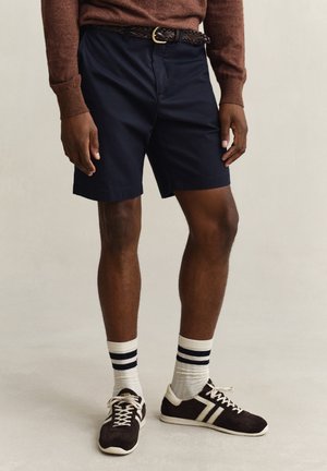Mann trägt marineblaue Shorts mit geflochtenem braunem Gürtel, braunen Pullover, weiße Socken mit schwarzen Streifen und braun-weiße Turnschuhe.