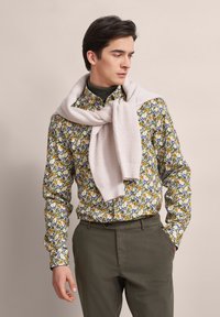 Blumengemusterte Hemd in Grün, Gelb und Blau; kombiniert mit einem cremefarbenen, gerippten Pullover, der um die Schultern gebunden ist; olivgrüne Hose.