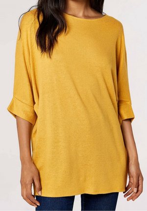 Femme portant un haut ample à manches trois-quarts couleur jaune moutarde, associée à un jean bleu foncé, sur un fond uni.