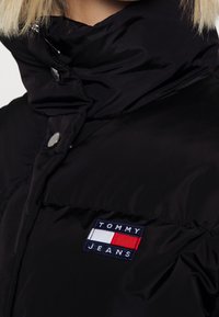Sort quiltet jakke med høj krave, med sølvfarvede trykknapper og en Tommy Jeans-logo patch i rød, hvid og blå.