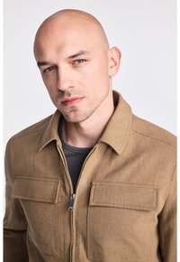 Brązowa marynarka z mieszanki lnu z zamkiem z przodu, dwoma kieszeniami na piersi i kołnierzykiem. Noszona na szarym koszuli o gładkiej fakturze.