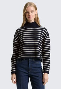 Pull en maille rayé bleu marine et crème avec un col montant, une coupe raccourcie et des poignets côtelés, associé à un jean denim bleu foncé.