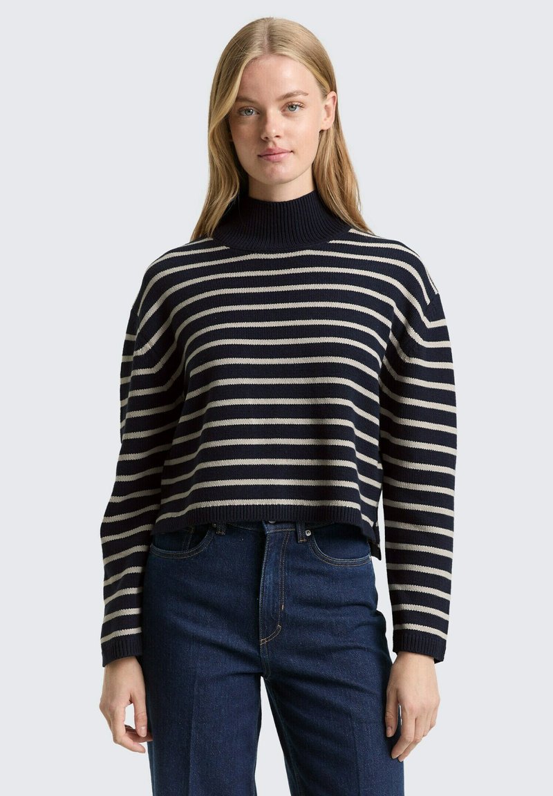 Pull en maille rayé bleu marine et crème avec un col montant, une coupe raccourcie et des poignets côtelés, associé à un jean denim bleu foncé.