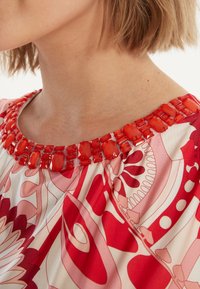 Rode jurk met een samengetrokken halslijn en een geborduurde kraag in verschillende tinten rood, op een gestreepte stof in felle rode en roze tinten.