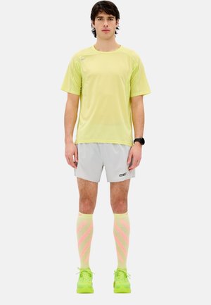 Helles gelbes Sportshirt, graue Shorts, knielange Socken mit pinken Streifen und grüne Sneakers. Das Material wirkt atmungsaktiv und passgenau.