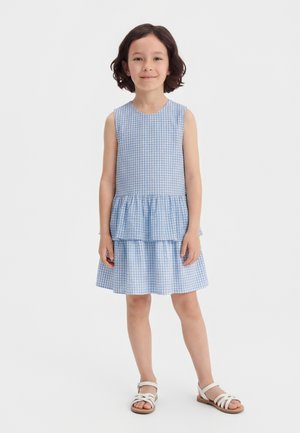 Νεαρή κοπέλα στέκεται, φοράει αμάνικο μπλε και άσπρο καρό φόρεμα με βολάν στη φούστα και λευκά σανδάλια.