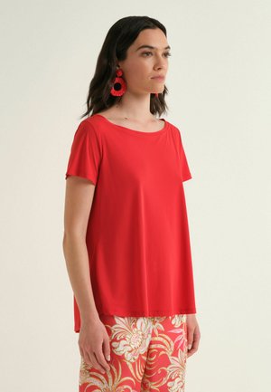 Donna che indossa una maglietta a maniche corte di un rosso brillante, grandi orecchini a cerchio rossi e pantaloni a fantasia rossa e bianca, in piedi davanti a uno sfondo semplice.