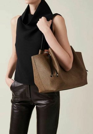 Borsa tote in pelle marrone con texture, con cordini neri, tenuta in una mano, abbinata a una canotta nera senza maniche e pantaloni in pelle marrone.