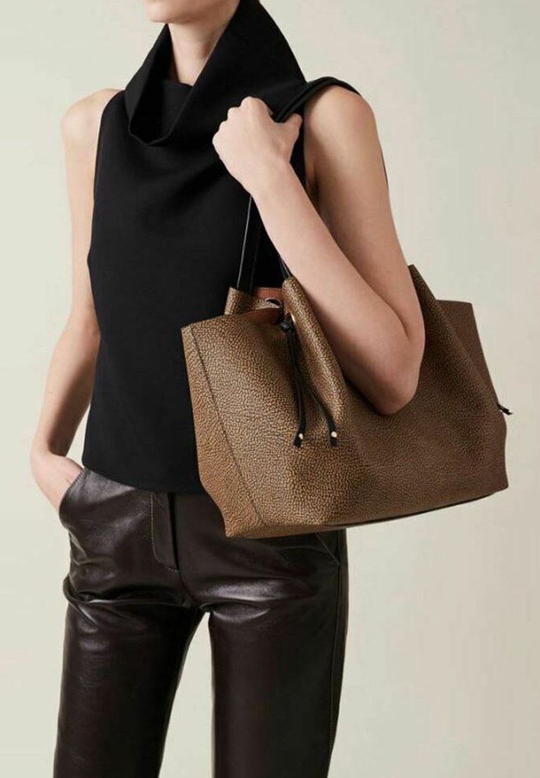 Borsa tote in pelle marrone con texture, con cordini neri, tenuta in una mano, abbinata a una canotta nera senza maniche e pantaloni in pelle marrone.