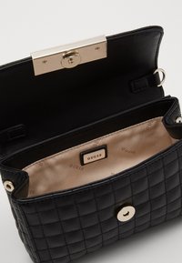 Czarna pikowana torebka na ramię z złotym zapięciem, beżowym wnętrzem i widoczną metką z logo. Z krótkim paskiem i detalem w postaci guzika.