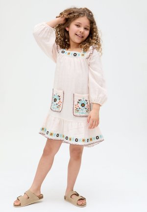 Young girl smiling, wearing a light pink dress with floral embroidery on pockets, hem, and neckline, posing with one hand in her curly hair.