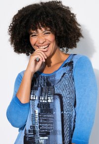 Chemise à manches longues bleue avec imprimé graphique texturé présentant un texte audacieux et des motifs. Tissu doux, col rond et manches contrastantes.