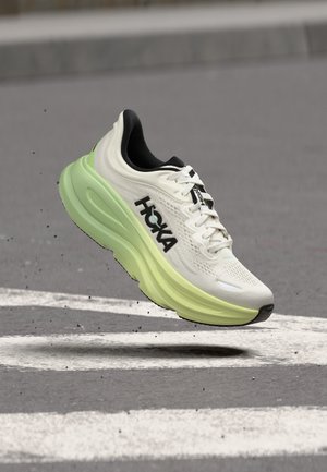 Chaussure de course Hoka blanche et vert néon en suspension au-dessus de l'asphalte avec des marquages routiers blancs et des détails noirs sur la semelle.