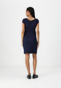 Vestido azul-marinho com mangas em pérola e detalhes franzidos nas costas. Ajustado ao corpo, até ao joelho, feito de um tecido suave. Usado com sapatos pretos.