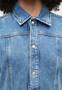 Ανοιχτό μπλε τζιν πουκάμισο με γιακά, που διαθέτει ασημί κουμπιά snap και ορατές ραφές. Το ύφασμα έχει υφή και κλασικό μοτίβο τζιν.