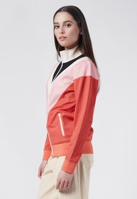 Färgglad zip-up jacka med en vit krage, med paneler i korall, rosa och svart, samt en sidoficka med en dold dragkedja.