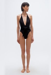 Γυναίκα που στέκεται ξυπόλυτη με μαύρο ολόσωμο μαγιό με τιράντες σε στυλ halter, βαθύ V ντεκολτέ και κορδόνι στη μέση, κοιτάζοντας προς τα αριστερά της, με φόντο απλό.