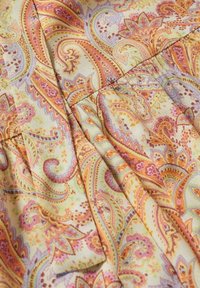 Tissu avec motif paisley détaillé dans des tons chauds d'orange, rose, jaune et beige, montrant des plis et des lignes de couture.
