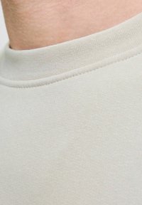 Lys grå stof, der viser en nærbillede af en rund hals med en blød, glat tekstur og subtile ribbede detaljer langs kraven.