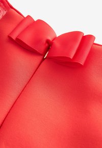 Tissu rouge avec un détail de nœud en ruban rouge texturé maintenu par une petite boucle dorée sur une surface lisse.
