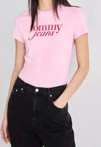 Camiseta de algodón rosa con mangas cortas que presenta "tommy jeans" en un guion rojo. Combinada con jeans de mezclilla negros de cintura alta.