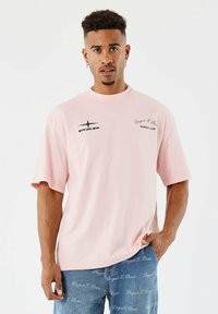 Hombre con cabello corto y rizado, vistiendo una camiseta estampada de un rosa claro y jeans azules con un patrón de escritura blanca, de pie frente a un fondo blanco.