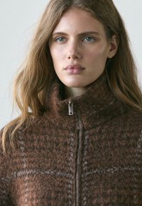 Brązowa dzianinowa kurtka z wysokim kołnierzem, z subtelnym wzorem w kratę i srebrnym zamkiem błyskawicznym. Miękka faktura i ciepły wygląd.