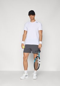 Valge sportlik T-särk, hallid lühikesed püksid, millel on tekst "BORG", valged spordijalatsid, hoiab tennisepalli ja reketit, must müts. Lihtne disain, vabalt langev lõige.