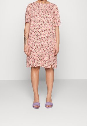 Personne portant une robe fleurie rose et blanche à manches courtes et longueur genoux, des sandales tissées violettes, un collier de perles, et un tatouage visible sur le bras.