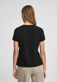 Camiseta negra de manga corta con cuello redondo, textura suave y corte relajado. El modelo lleva pantalones beige, mostrando la vista trasera de la camiseta.