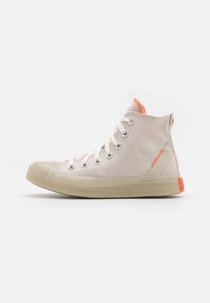Beige canvas hoge sneaker met witte veters, doorschijnende beige zool en oranje stikseldetails op de tong en hielstuk.